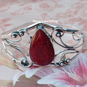 May include: A silver bangle bracelet with a large, teardrop-shaped red gemstone set in the center. The bracelet has a delicate, ornate design.