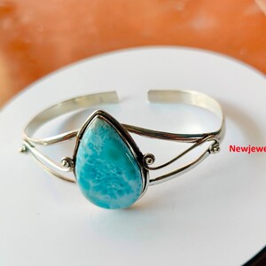 May include: A silver cuff bracelet with a pear-shaped, light blue larimar stone. The stone is set in a silver bezel and the bracelet has a simple, open design. The jewellery is displayed on a white surface.