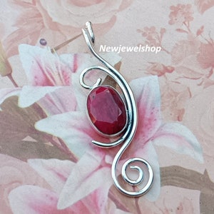 May include: A silver pendant with a red gemstone set in a swirl design. The pendant is on a white background with pink flowers.
