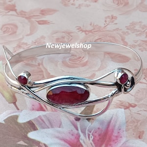 May include: A silver bangle bracelet with a delicate, intricate design featuring three red gemstones. The bracelet is open and adjustable.