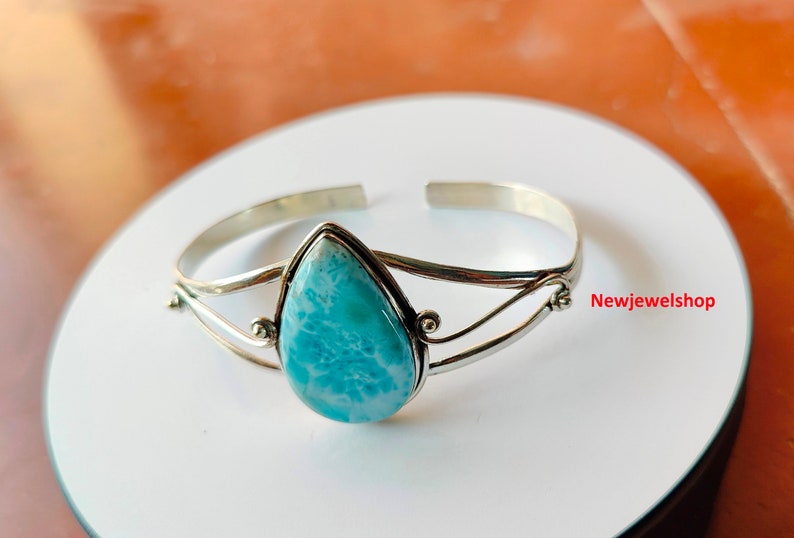 May include: A silver cuff bracelet with a pear-shaped, light blue larimar stone. The stone is set in a silver bezel and the bracelet has a simple, open design. The jewellery is displayed on a white surface.