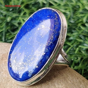 May include: A silver ring featuring a large, oval-shaped lapis lazuli stone. The deep blue stone has flecks of gold and is set in a polished silver band. The ring is displayed on a natural stone surface against a green background.