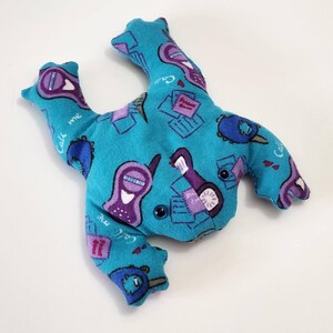 May include: A turquoise plush frog toy with a repeating pattern of purple flip phones, notepads, and the words "Call me." The frog has two black button eyes and is made of soft fabric.