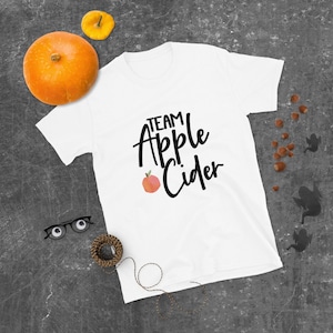 May include: White t-shirt with the text "Team Apple Cider" in black script font. A red apple with a green leaf is next to the text.