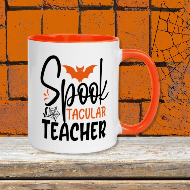 Puede incluir: Taza de caf&eacute; blanca con borde y asa naranja. La taza tiene un gr&aacute;fico negro que dice "Spooktacular Teacher" con un dise&ntilde;o de murci&eacute;lago y telara&ntilde;a.