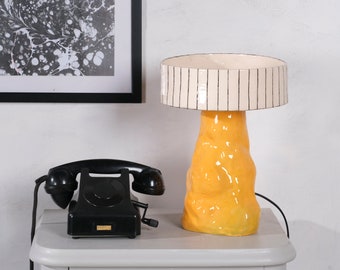 May include: A yellow ceramic table lamp with a white striped shade. The lamp is on a white table next to a black rotary phone.