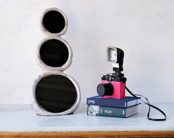 May include: A decorative, three-circle mirror with a textured, off-white frame stands next to a pink camera with a black lens and flash. The camera rests on two books with the titles "Diana F" and "Film". A black strap extends from the camera.