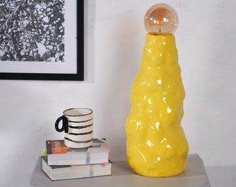 May include: A yellow ceramic table lamp with a textured surface and a round light bulb. The lamp is sitting on a gray table.