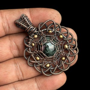 Puede incluir: Un colgante artesanal con una piedra preciosa verde oscuro en el centro, encerrada en intrincados alambres de cobre. El colgante tiene un dise&ntilde;o floral con peque&ntilde;os acentos dorados. La argolla tambi&eacute;n es de alambre de cobre.
