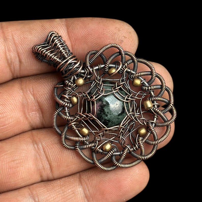 Puede incluir: Un colgante artesanal con una piedra preciosa verde oscuro en el centro, encerrada en intrincados alambres de cobre. El colgante tiene un dise&ntilde;o floral con peque&ntilde;os acentos dorados. La argolla tambi&eacute;n es de alambre de cobre.