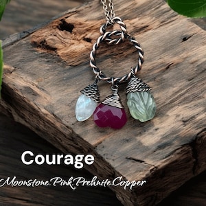 Könnte beinhalten: Eine silberne Halskette mit einem verdrehten, kreisförmigen Anhänger, der drei Edelsteinanhänger hält. Die Anhänger umfassen einen weißen Mondstein, einen facettierten rosa Stein und einen grünen Prehnit. Das Wort "Courage" ist in weißer Schrift geschrieben.