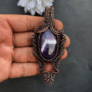 Pode incluir: Pingente artesanal de fio de cobre com uma pedra preciosa oval polida, roxa e branca. O intrincado trabalho de arame envolve a pedra, criando um design ornamentado. O pingente &eacute; suspenso por uma argola de arame tran&ccedil;ado.