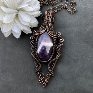 Pode incluir: Um colar com pingente artesanal com uma grande pedra de ametista oval. A pedra roxa est&aacute; emoldurada em uma estrutura intrincada, enrolada em fio de metal cor de cobre. O pingente pende de uma corrente combinando, mostrando um design art&iacute;stico &uacute;nico.