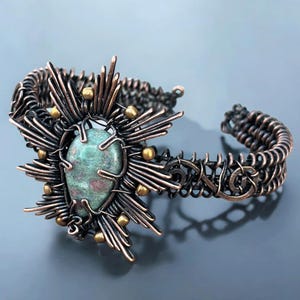May include: A handcrafted copper bracelet featuring a central, oval-shaped green stone with red inclusions. The stone is surrounded by a sunburst design with gold-colored accents. The bracelet has a woven chain design.