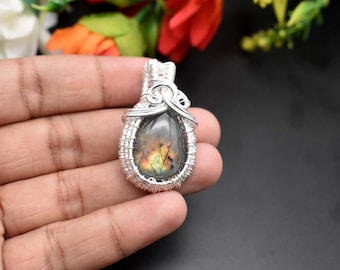 May include: Silver wire wrapped pendant featuring a iridescent labradorite gemstone. The stone has a dark gray base with flashes of green and gold.
