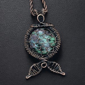 May include: A handcrafted copper wire pendant featuring a round, green and purple stone. The stone is encased in a woven wire cage, with decorative wire detailing and a stylized tail design. The pendant hangs from a matching copper chain.