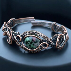 May include: A copper wire bracelet with an oval stone at its centre. The stone displays green and red colours. The wire is intricately woven around the stone and forms the bracelet's structure. The bracelet is a unique piece of jewellery.