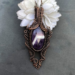 Pode incluir: Colar com pingente artesanal de arame de cobre com uma pedra preciosa de ametista oval. A ametista &eacute; roxa escura com estrias brancas. O pingente tem um design intrincado de arame tecido e pende de uma corrente delicada.