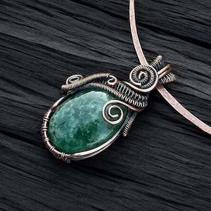 Può includere: Un ciondolo di pietra verde con avvolgimento di filo di rame. Il ciondolo è di forma ovale e ha un colore verde scuro con macchie bianche. Il filo di rame è attorcigliato e avvolto attorno alla pietra, creando un design decorativo.