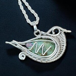 Peut inclure: Un pendentif en fil métallique argenté en forme d'oiseau, doté d'une pierre texturée verte et rouge violacé en son centre. Le pendentif est suspendu à une chaîne torsadée argentée assortie et comporte une petite perle noire pour l'œil.