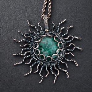 Könnte beinhalten: Eine handgefertigte Anhänger-Halskette mit Sonnenmotiv. Der Anhänger hat einen zentralen, runden, grünen Stein, der von einem silberfarbenen Drahtrahmen umgeben ist. Die Sonnenstrahlen bestehen aus gedrehtem Draht, was ein einzigartiges Kunstwerk ergibt.