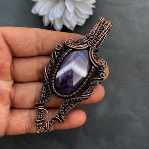 Pode incluir: Um pingente artesanal com uma pedra preciosa de ametista oval polida. A pedra roxa &eacute; colocada em uma moldura de arame de cobre, intrincadamente tecida e moldada. O pingente tem um design art&iacute;stico &uacute;nico, para joias.