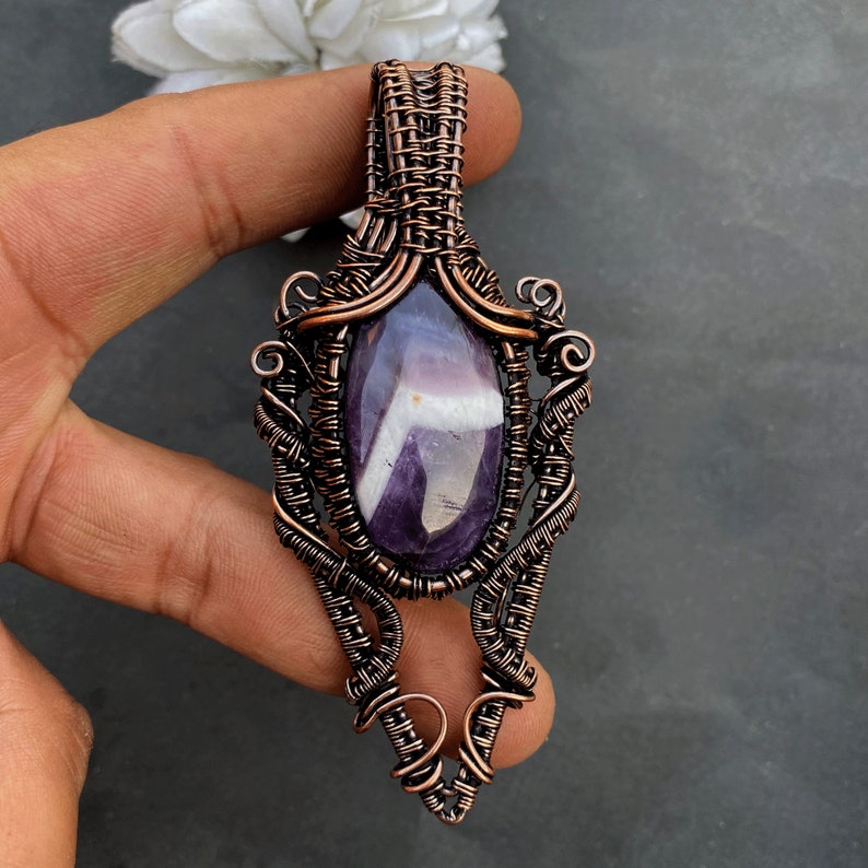 Pode incluir: Um pingente artesanal com uma pedra de ametista polida em forma oval com estrias brancas, envolta em uma intrincada tecelagem de fio de cobre. O pingente tem formato de gota com redemoinhos decorativos e uma argola para uma corrente. A ametista &eacute; de cor roxa escura.