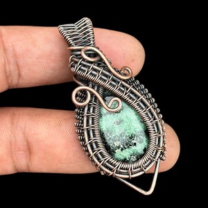 May include: A handcrafted pendant featuring a teardrop-shaped green stone encased in intricate copper wire. The wirework includes decorative swirls and a triangular base, creating a unique and artistic jewelry piece.