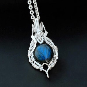 May include: A silver pendant necklace featuring a round, blue gemstone. The gemstone is encased in a detailed wire-wrapped setting. The pendant hangs from a delicate silver chain, creating an elegant and eye-catching piece of jewelry.