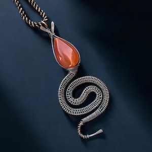 Puede incluir: Un collar con colgante artesanal con una piedra preciosa naranja en forma de lágrima engastada en metal plateado. El colgante está unido a un diseño de alambre enrollado, que se asemeja a una serpiente, y cuelga de una delicada cadena.