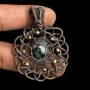 May include: A handcrafted pendant featuring a dark green gemstone at the center, encased in intricate copper wirework. The wire is woven into a decorative pattern with small gold-colored beads. The pendant is held against a black background.