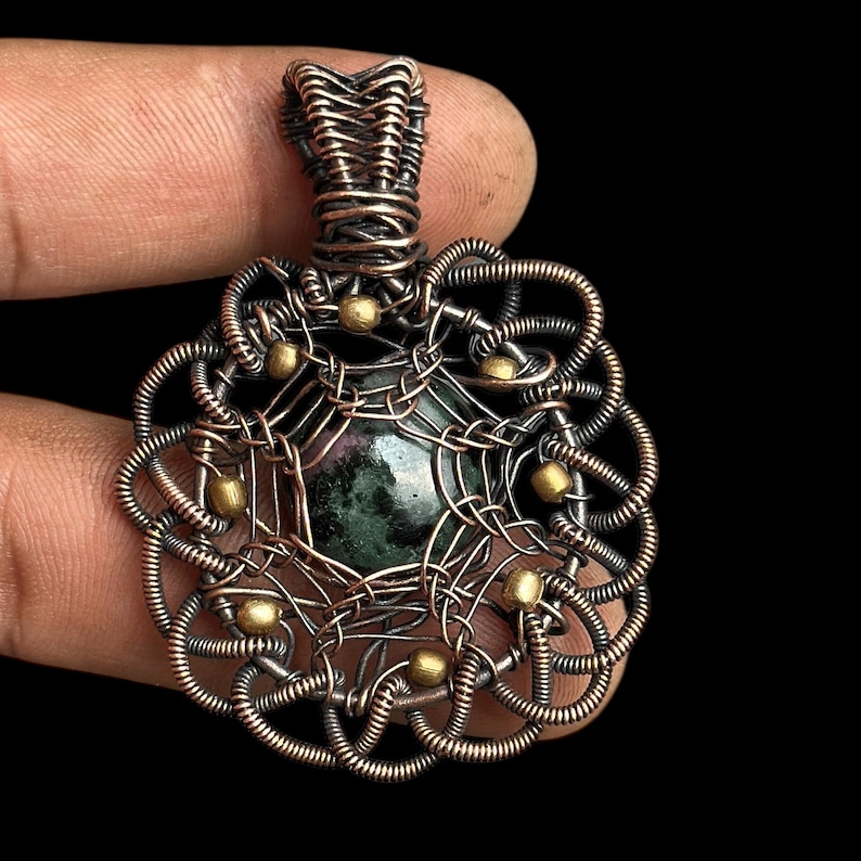 Puede incluir: Un colgante artesanal con una piedra preciosa verde oscuro en el centro, encerrada en un intrincado trabajo de alambre de cobre. El alambre est&aacute; tejido en un patr&oacute;n decorativo con peque&ntilde;as cuentas doradas. El colgante se muestra sobre un fondo negro.