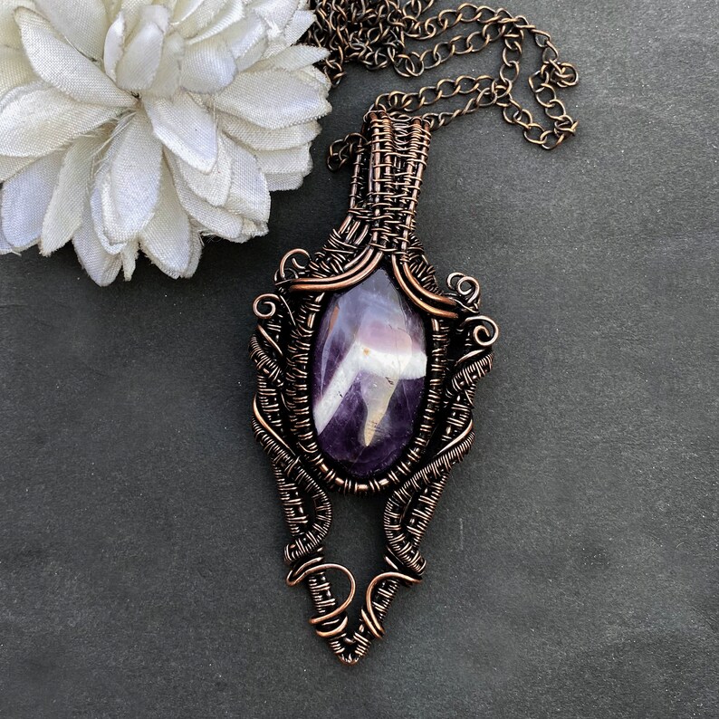 Pode incluir: Colar com pingente artesanal de fio de cobre com uma pedra de ametista oval. O pingente tem um fio intrincado e um design decorativo. A corrente &eacute; de cor cobre. O colar &eacute; exibido em uma superf&iacute;cie cinza escuro.