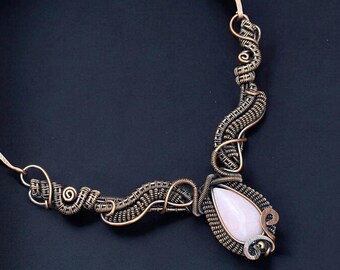 May include: A handcrafted copper wire necklace with a teardrop-shaped, pale pink gemstone pendant. The wire is intricately woven and coiled, creating a unique, artistic design. The necklace is displayed against a dark background.