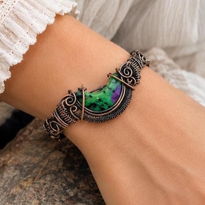 May include: A wire-wrapped bracelet featuring a crescent-shaped green and purple zoisite gemstone. The band is constructed from dark copper-toned metal wire, intricately coiled and woven into decorative swirls surrounding the central stone.