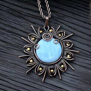 May include: A copper wire necklace with a blue glass pendant in the shape of a sun. The pendant is surrounded by a wire design with gold beads.