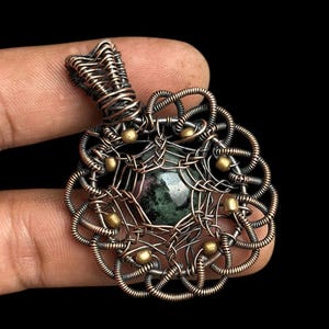 Puede incluir: Un colgante artesanal con una piedra central de color verde oscuro, rodeada de intrincados trabajos de alambre de cobre. Peque&ntilde;as cuentas doradas est&aacute;n espaciadas uniformemente alrededor del dise&ntilde;o. La argolla del colgante tambi&eacute;n es de alambre de cobre trenzado.