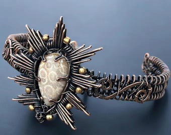 May include: Copper-coloured cuff bracelet with an oval fossil stone centrepiece. The stone is framed by a sunburst design, with small gold-coloured beads. The bracelet features detailed wirework.