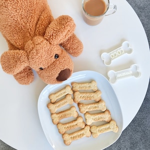 以下が含まれることがあります： 白い皿に、犬の骨の形をしたクッキーがいくつか置かれています。クッキーにはすべて「Plastic」という文字が押されており、皿の上に円形に並べられています。皿の隣には、白い表面に茶色のぬいぐるみ犬が置かれています。背景には、茶色の液体の入ったグラスがあります。