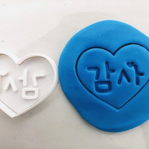 Peut inclure: Un emporte-pièce en plastique blanc en forme de cœur avec le mot coréen "사랑" (signifiant "amour") à l'intérieur. L'emporte-pièce est utilisé pour créer une empreinte en forme de cœur dans de l'argile à modeler bleue.