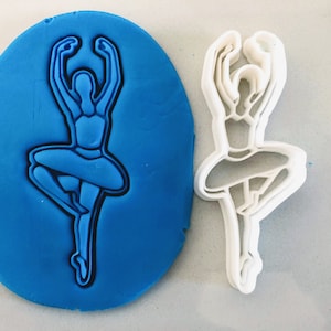 May include: A white plastic cookie cutter in the shape of a ballerina. The cutter is shown next to a blue fondant impression of the cutter.