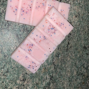 May include: Two pink wax melts with a glittery finish. The wax melts are divided into sections and have a speckled appearance with blue and red glitter.