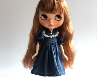 May include: A doll with long, wavy, auburn hair wearing a dark blue dress with a white lace collar and pearl buttons. The doll is wearing black shoes with a white bow.