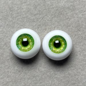 May include: A pair of green glass doll eyes with a white iris and a black pupil. The eyes are set in a white plastic base.