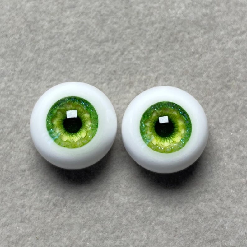 May include: A pair of green glass doll eyes with a white iris and a black pupil. The eyes are set in a white plastic base.