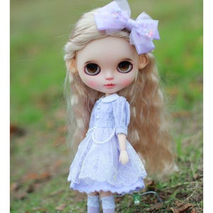 May include: A doll with blonde hair wearing a lavender dress with lace detailing and a pearl necklace. The doll has brown eyes and is wearing a lavender bow in her hair.