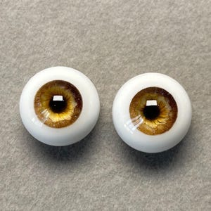 May include: Two brown and gold doll eyes with a black pupil. The eyes are set in a white plastic base.