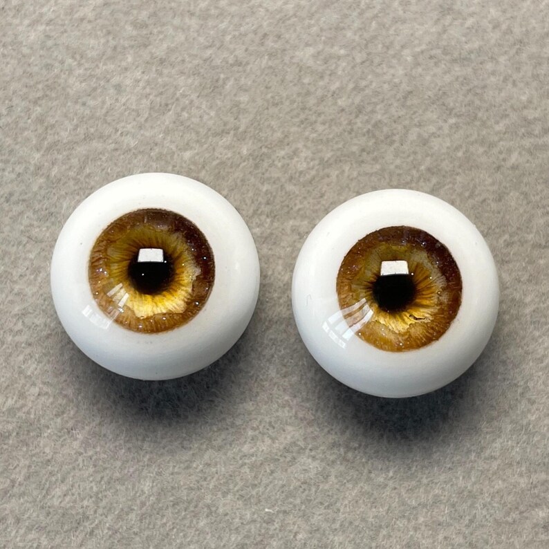 May include: Two brown and gold doll eyes with a black pupil. The eyes are set in a white plastic base.