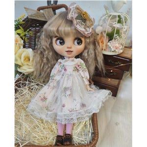 May include: A Blythe doll with long, wavy blonde hair, wearing a floral print dress with lace trim and a matching hat. The doll is posed in a wicker basket, with a decorative birdcage and wooden boxes in the background.