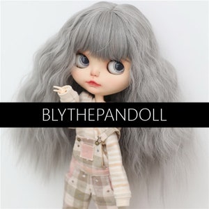 Könnte beinhalten: Eine Puppe mit langen, welligen, silbergrauen Haaren, die ein kariertes Latzkleid und ein gestreiftes Langarmshirt trägt. Die Puppe hat große, blaue Augen und eine hellrosa Nase. Der Text "BLYTHEPANDOLL" ist im Bild sichtbar.